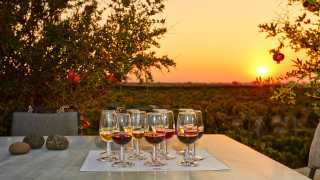 Glasses of volcanic wine on a table with the sun setting in the background over Santorini's vineyards